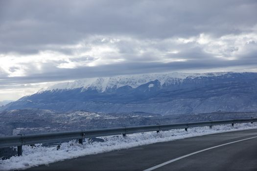 A winding road with snowy mountains under a cloudy sky creates a picturesque winter scene.