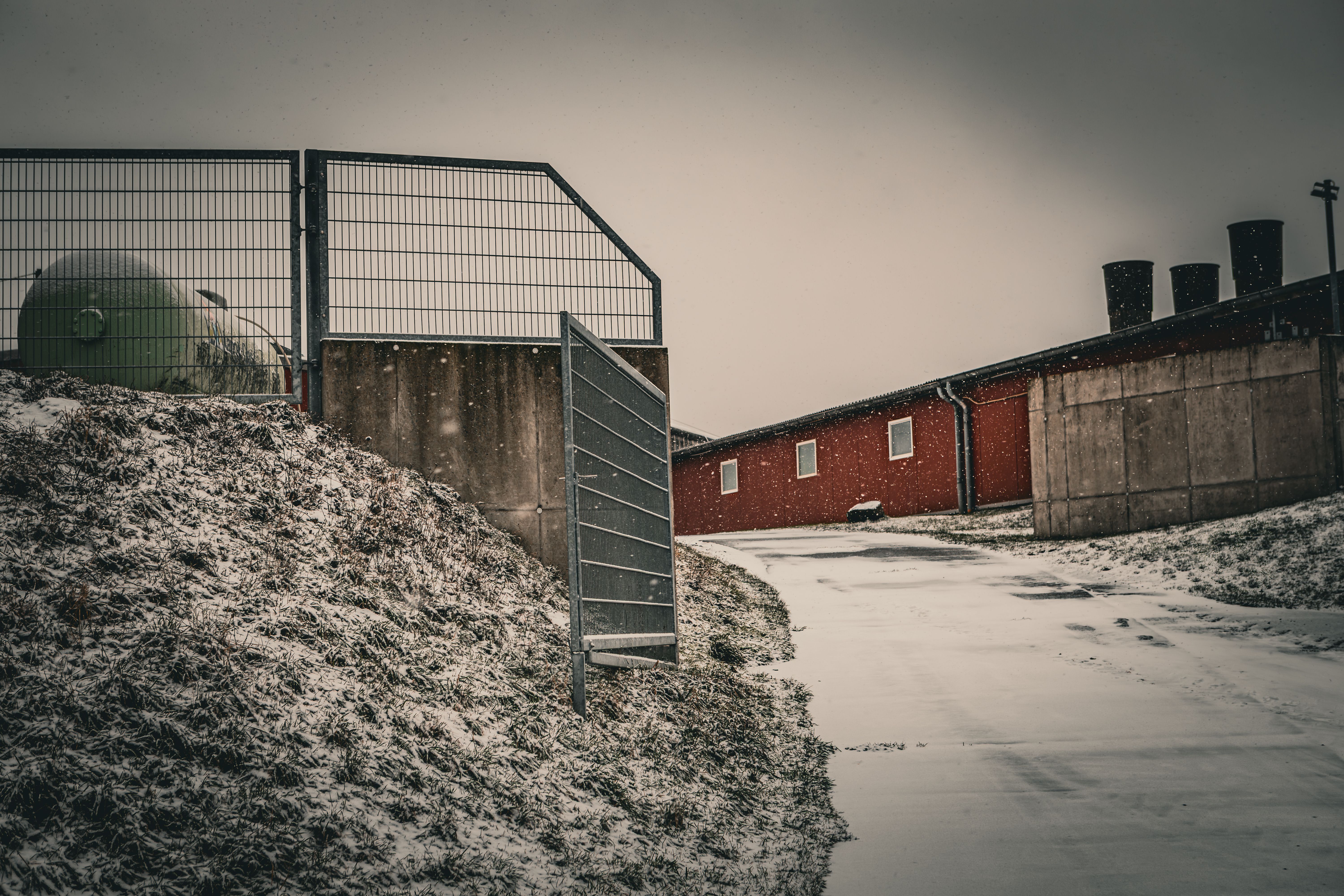 gratis Een besneeuwd industrielandschap met een metalen poort die toegang geeft tot een rood gebouw, onder winterse omstandigheden. Stockfoto