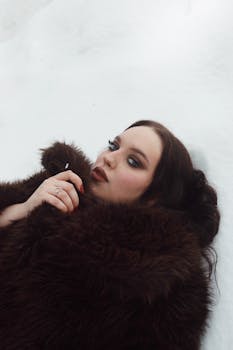 A stylish woman in a fur coat lies elegantly on the snow, gazing upward.