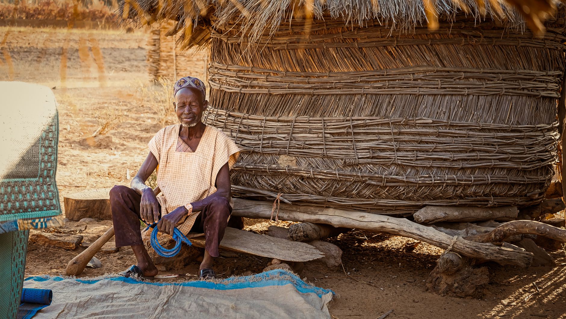 Elderly man relaxes outside a traditional woven hut on a sunny day.
