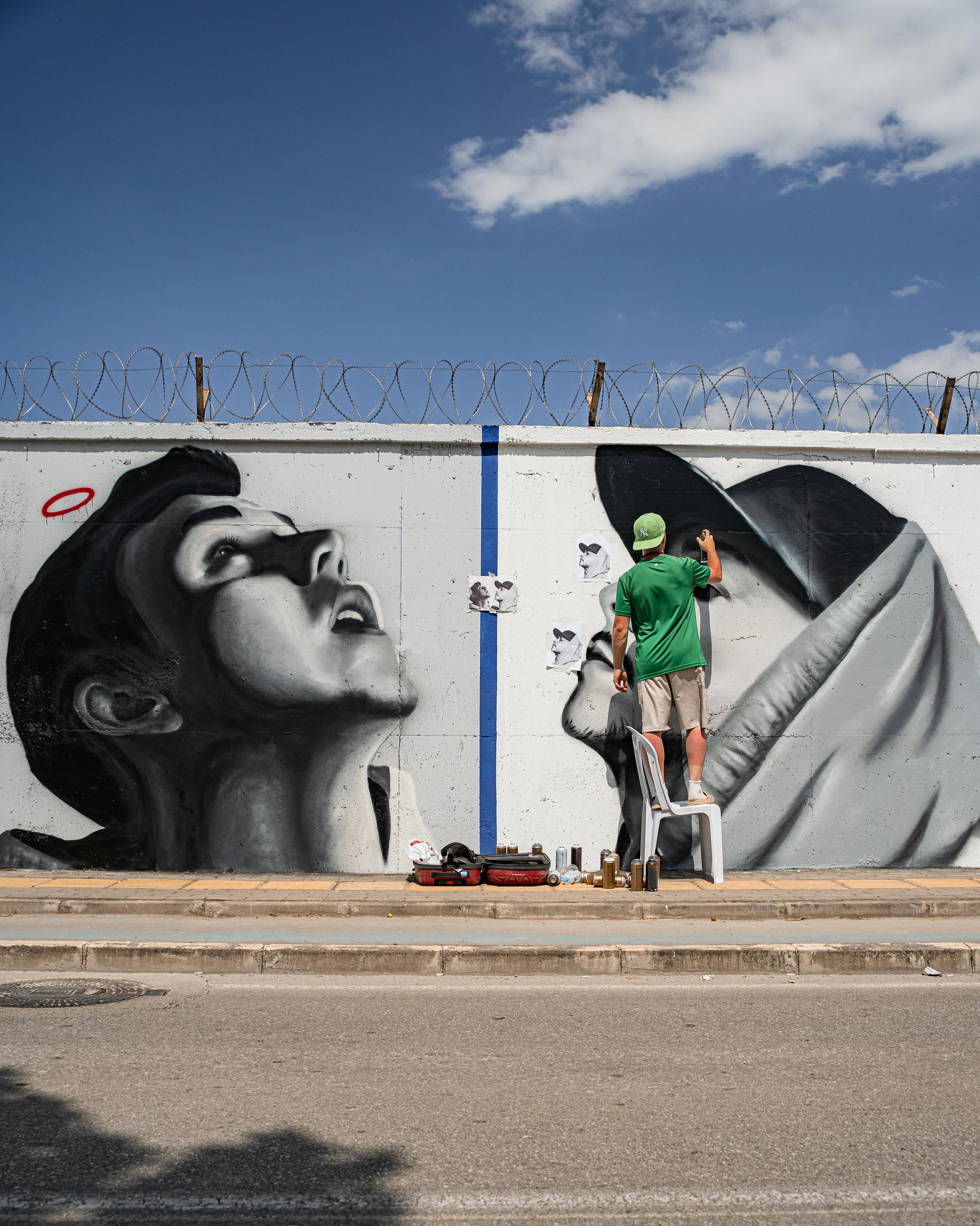 Street artist creating a black and white mural on an urban wall under a sunny sky.