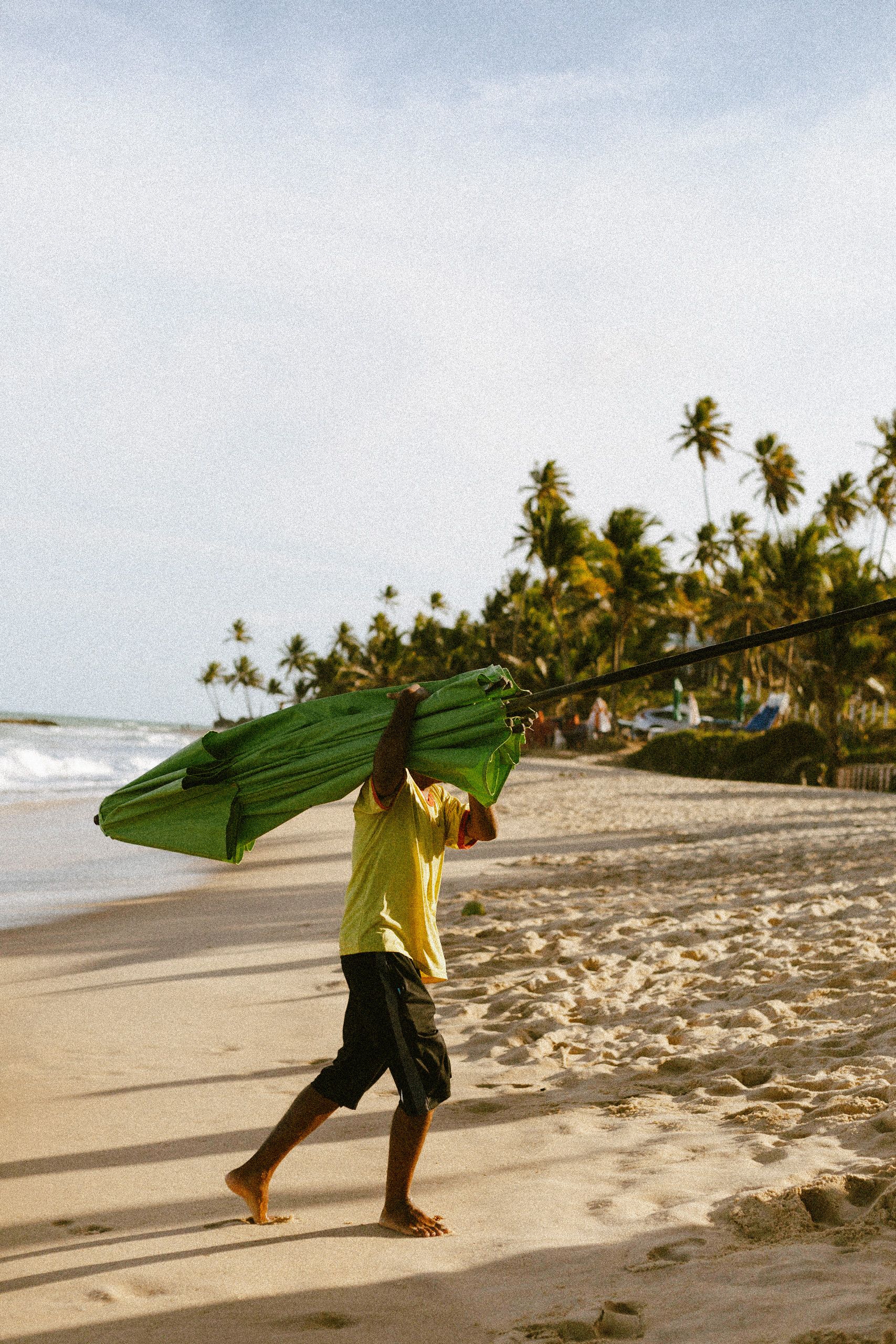 Man walking on João Pessoa beach, carrying a green umbrella during a sunny day.