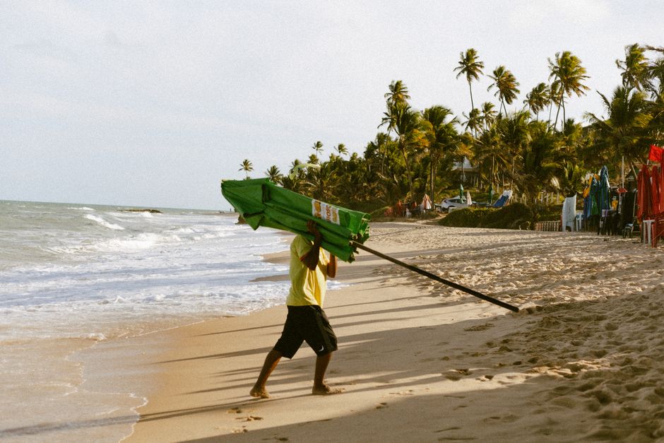 A beach vendor carrying an umbrella along a tropical shoreline in João Pessoa, Brazil.