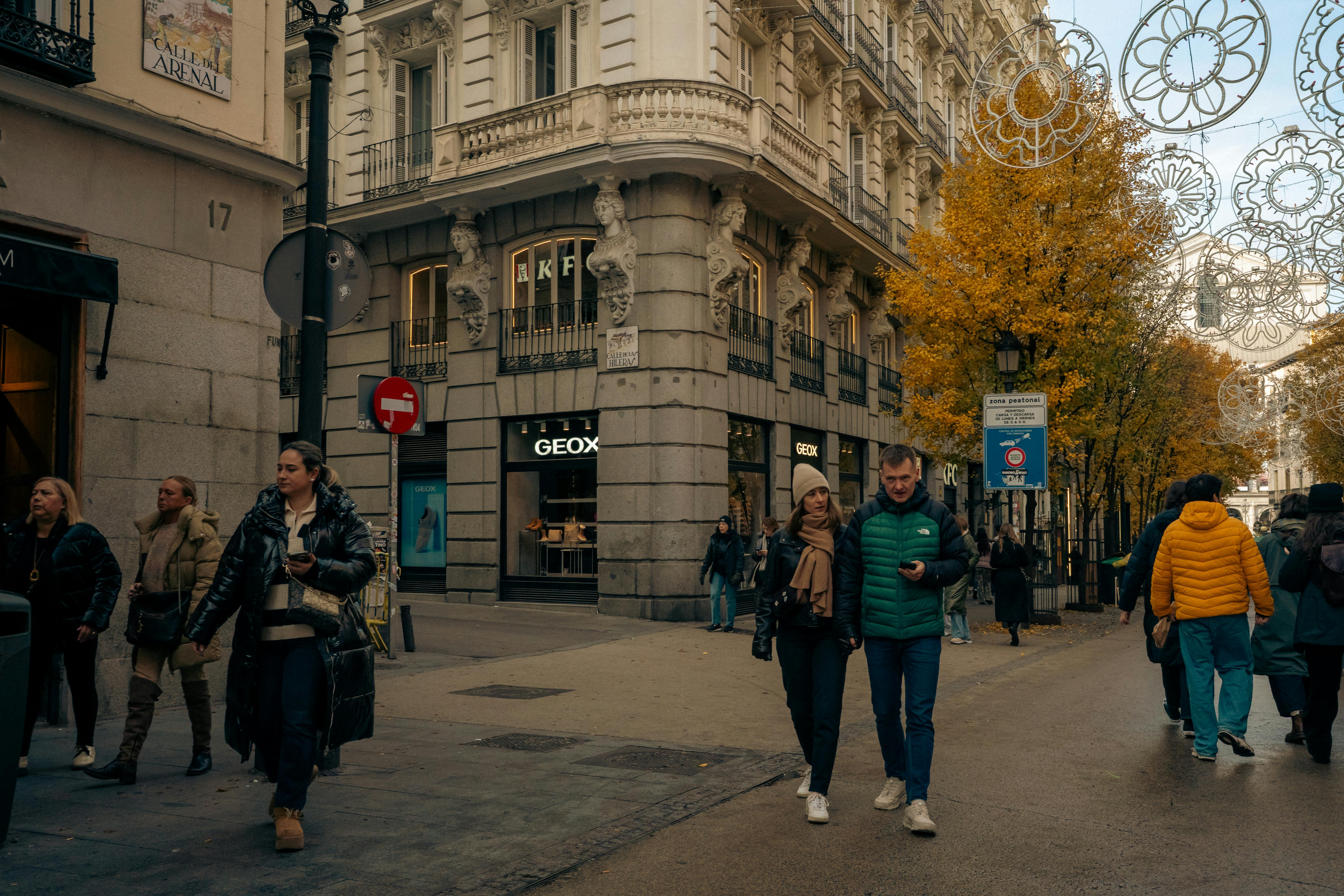 Street scene in Madrid's Calle del Arenal with autumn trees, shoppers, and European architecture.