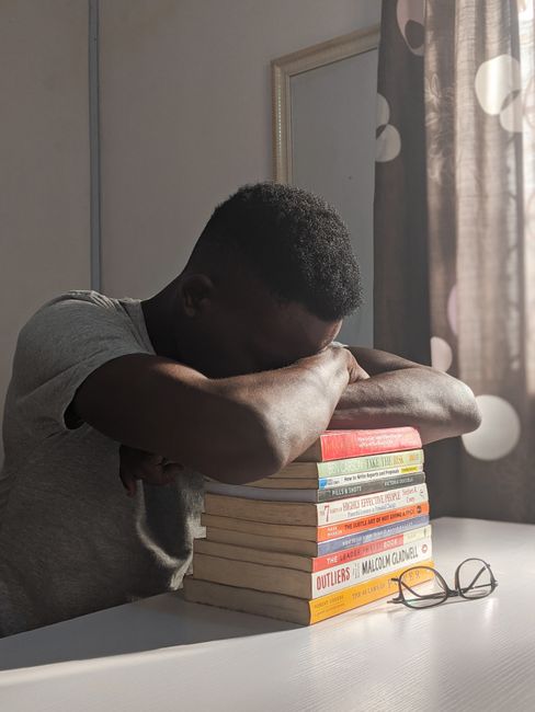 A young man rests on a stack of books at a desk, sunlight filtering through a window. A young man rests on a stack of books at a desk, sunlight filtering through a window.