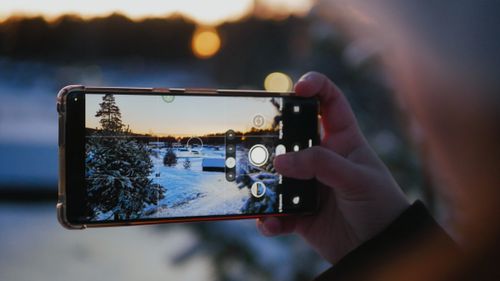 Free A hand holding a smartphone capturing a snowy landscape during sunset. Stock Photo