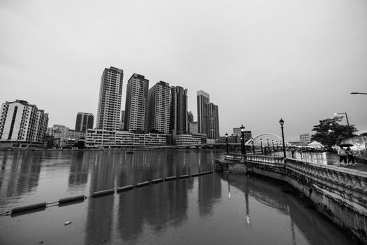 Black and white cityscape with tall buildings reflecting in a calm river during the day.