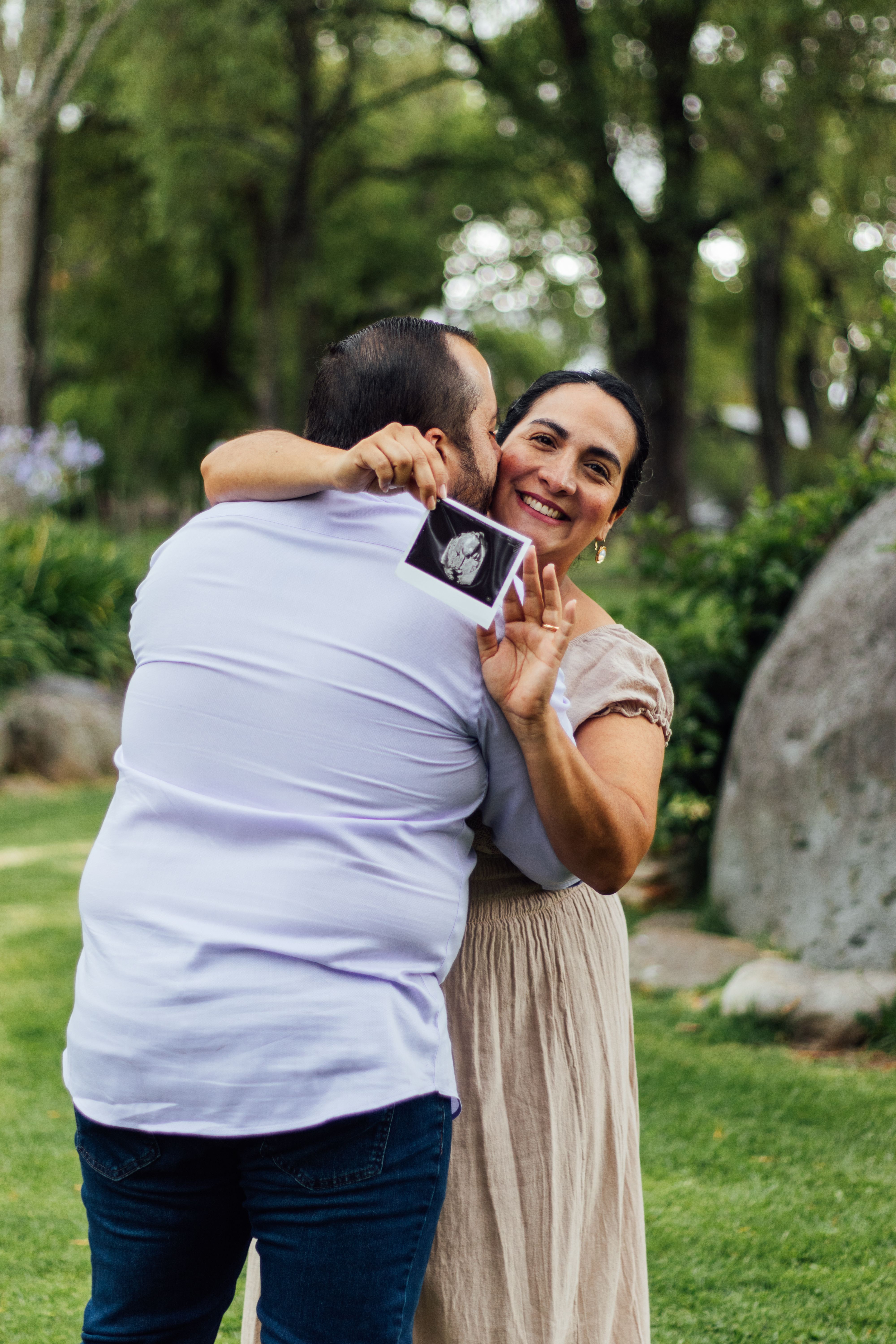 Expectant couple joyfully hugging outdoors, holding ultrasound photo.