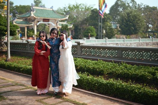 Three women in colorful traditional dresses pose with joy at an Asian pagoda.