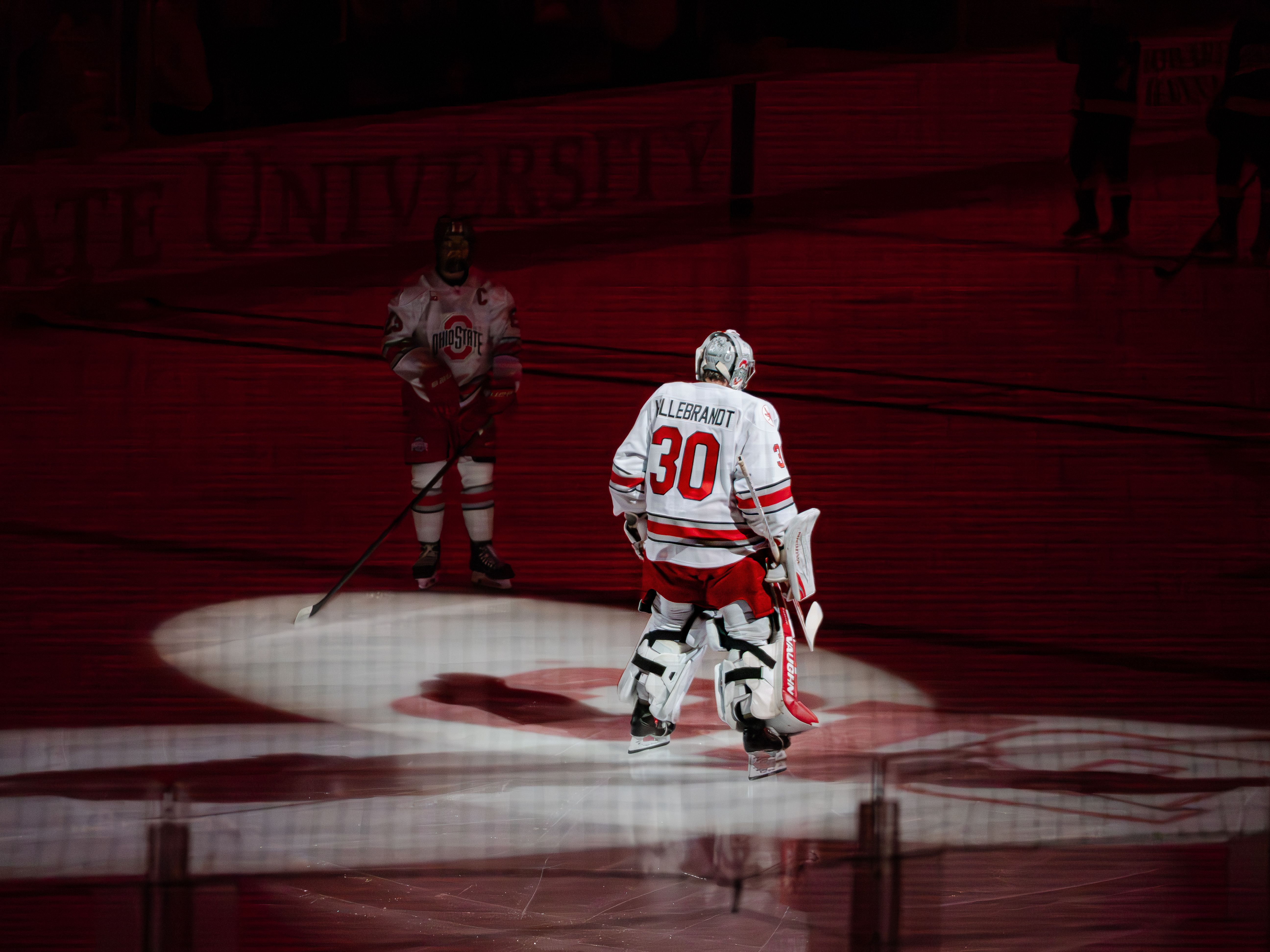 Ice hockey players in spotlight on a rink during a match under red lighting.