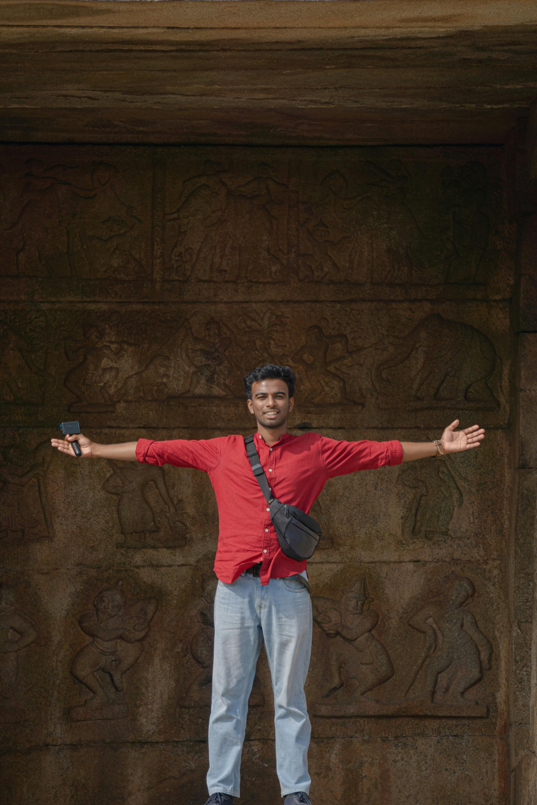 A man in a red shirt poses with outstretched arms in front of an intricately carved stone wall.