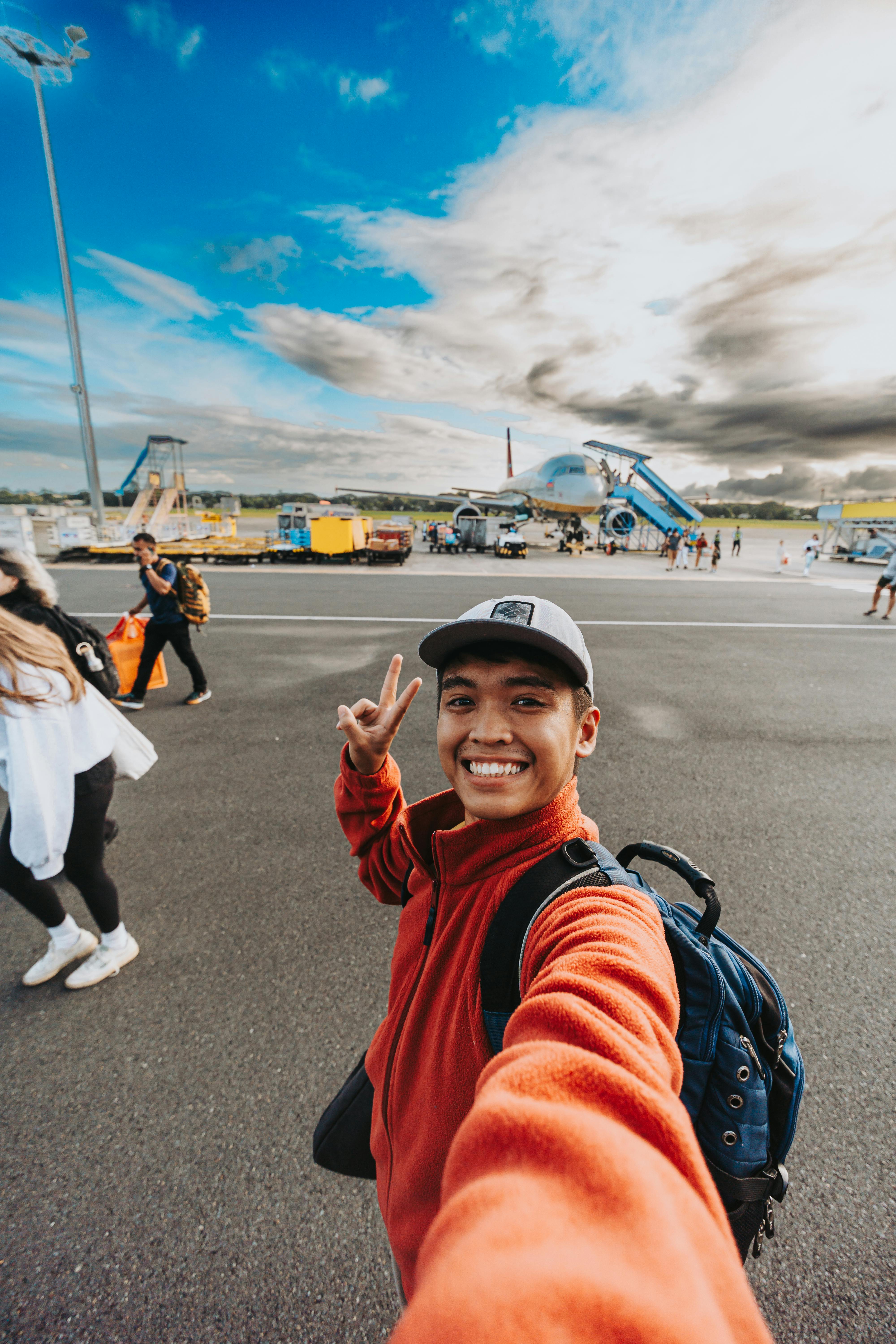 A cheerful young man takes a selfie on the airport tarmac with clear skies and airplanes in the background.