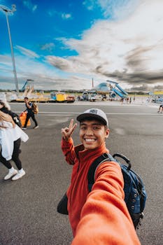 A cheerful young man takes a selfie on the airport tarmac with clear skies and airplanes in the background.