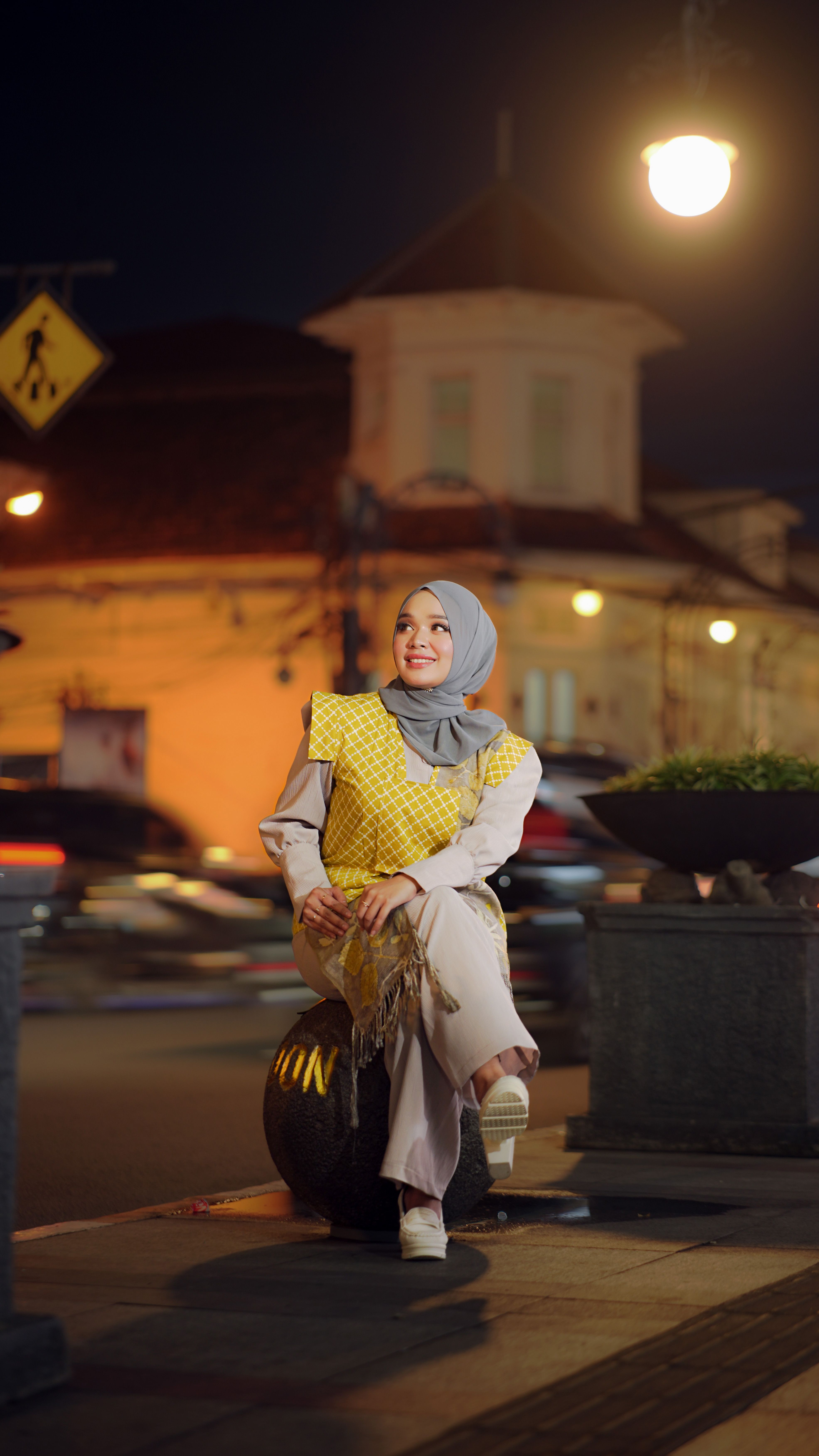 A fashionable woman sitting outdoors on a lit street at night in Bandung, Indonesia.