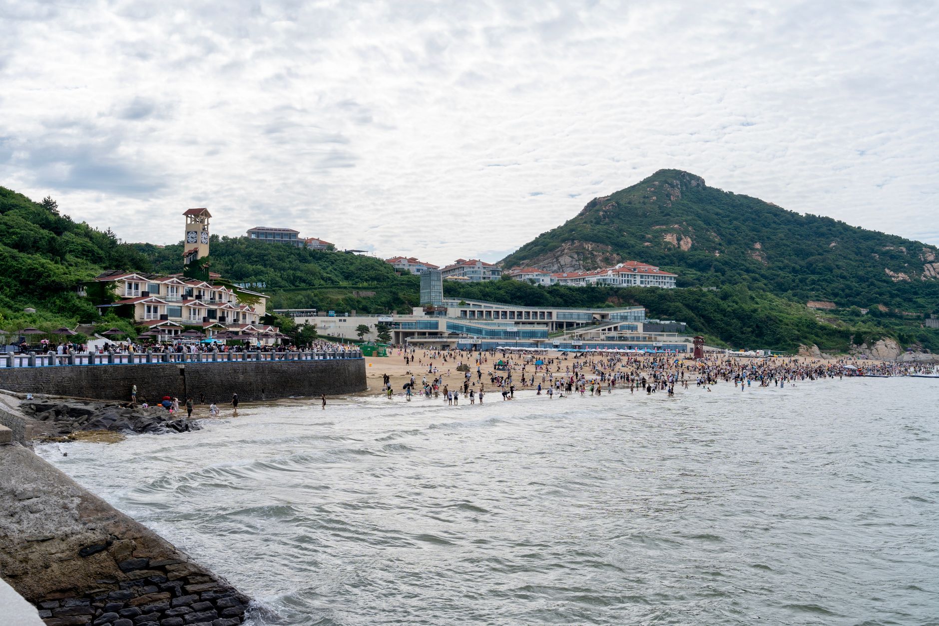 Bustling beachfront with people enjoying a summer day against a backdrop of lush hills and buildings.