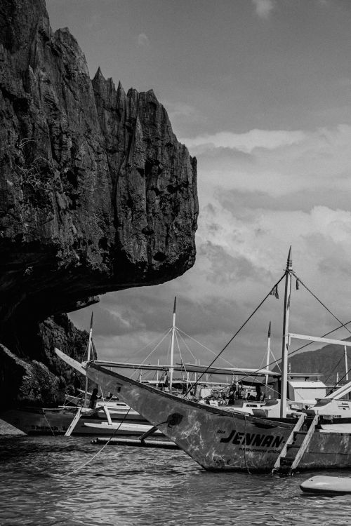 Free Monochrome scene of boats docked by a rugged rocky coastline under cloudy skies. Stock Photo