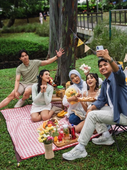 Free A group of friends having fun at a picnic in a Jakarta park, capturing joyful moments with a smartphone. Stock Photo