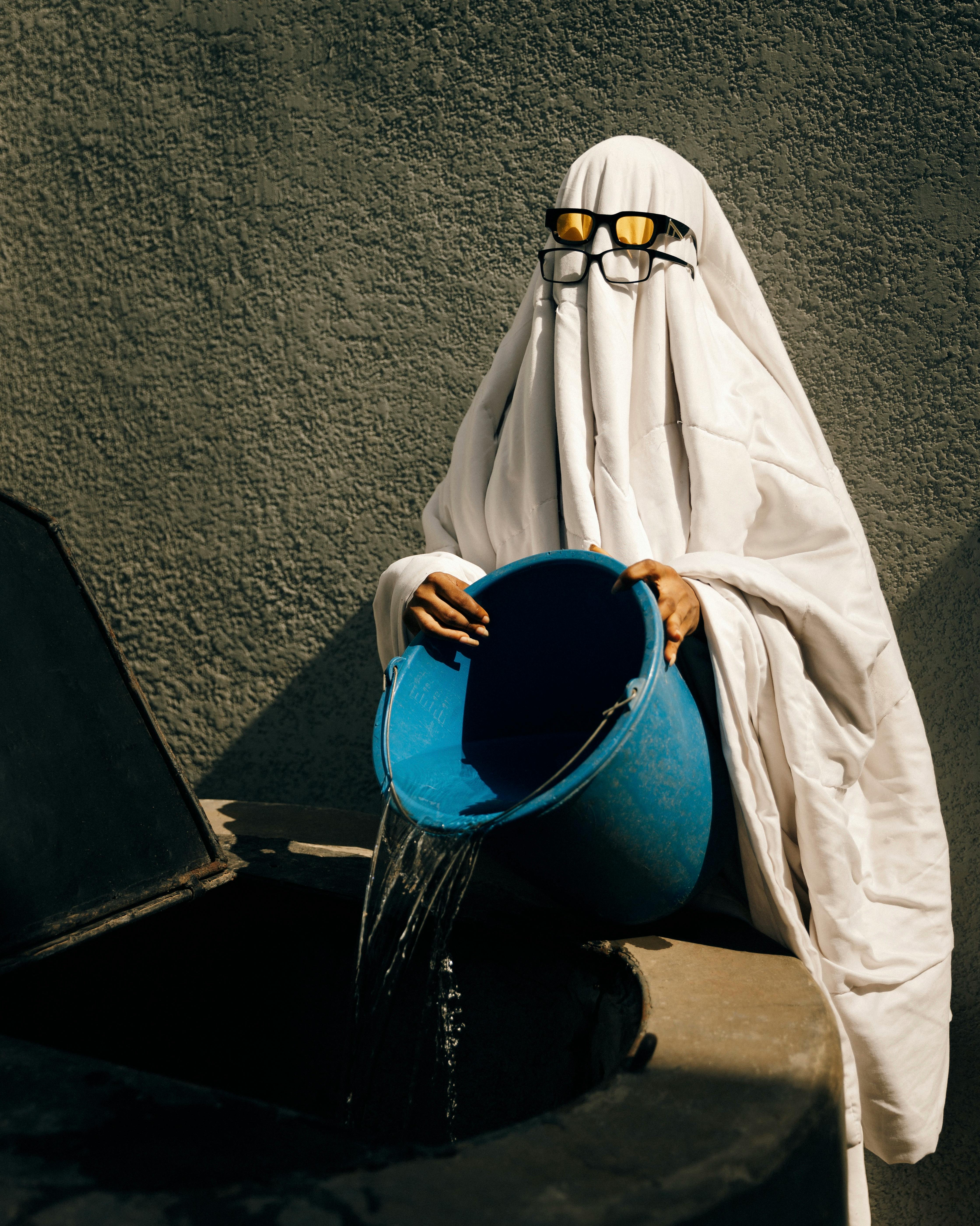 A person dressed as a ghost uses a blue bucket to pour water outdoors, creating a humorous scene.