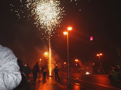 A lively city street with fireworks illuminating the night sky and people enjoying the show.