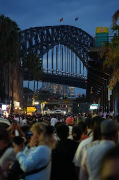 Crowd gathers near the iconic Sydney Harbour Bridge at dusk, creating a vibrant atmosphere.