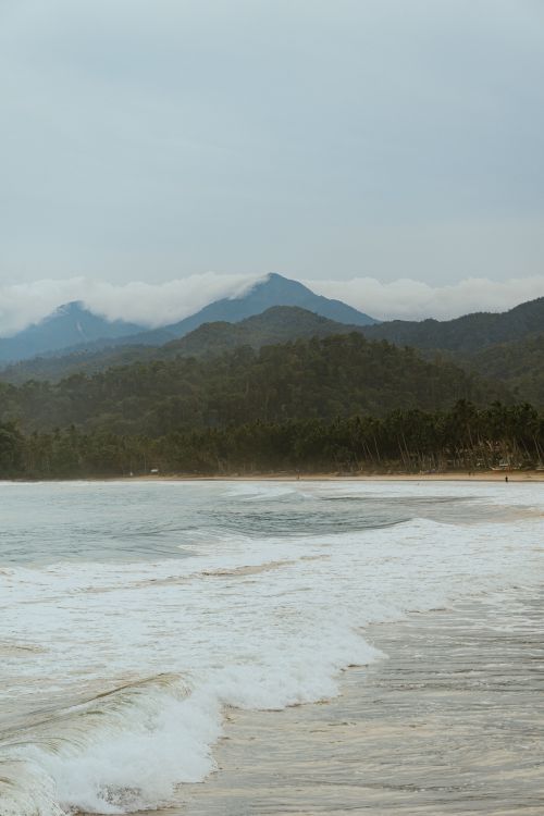 Free Serene beach scene with waves and distant mountains under a cloudy sky. Stock Photo