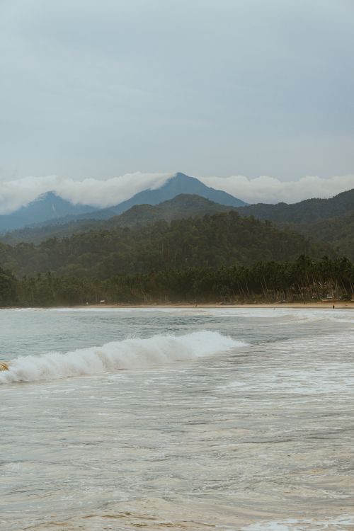 Free A serene beach scene with waves and mountains under cloudy skies. Stock Photo