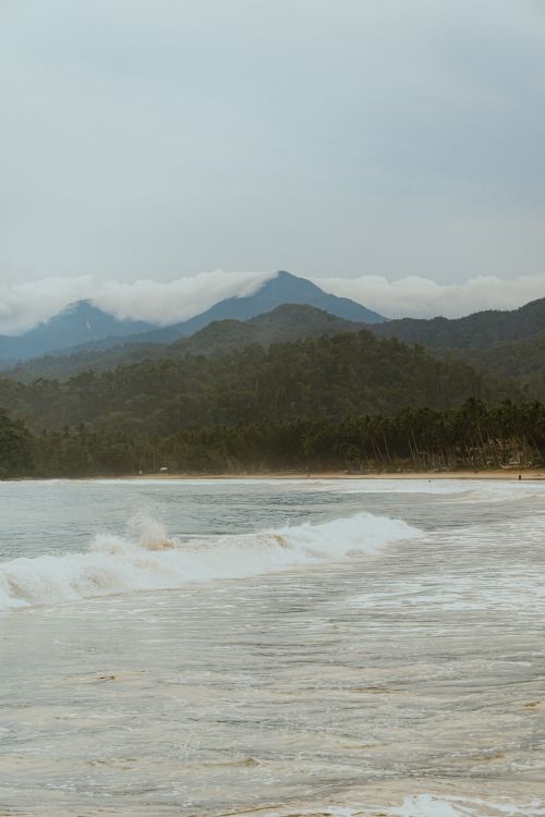 Free Breathtaking view of waves crashing on a beach with mountains in the background, creating a serene coastal landscape. Stock Photo