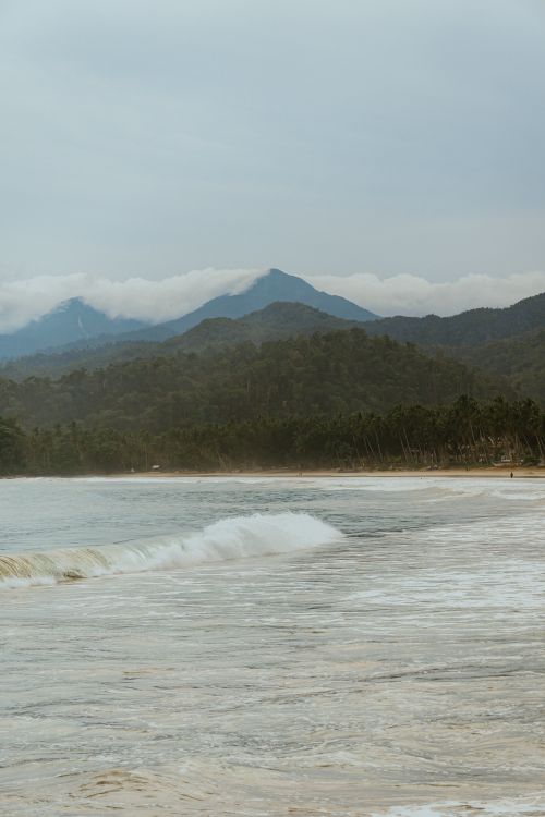 Free Serene tropical beach scene with gentle waves and mountain backdrop, ideal for travel and nature themes. Stock Photo