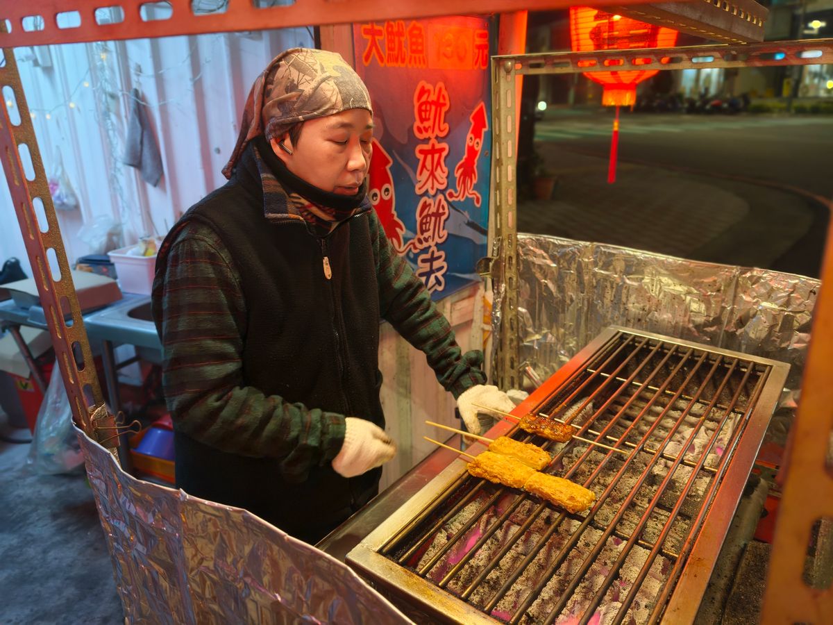 A vendor in a patterned headscarf and fleece grills squid skewers over a foil-lined charcoal cart at dusk, a red lantern glowing across the lane and a cartoon squid sign behind her.