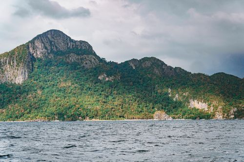 Free A picturesque mountain view over a serene ocean with lush, dense forest under a cloudy sky. Stock Photo