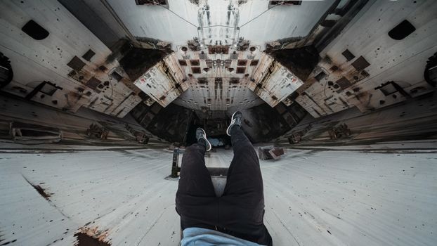 Man's legs dangle over edge of tall, rusted, industrial structure.
