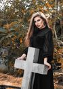 Serious woman with long hair wearing black dress standing near cross shaped tombstone in cemetery
