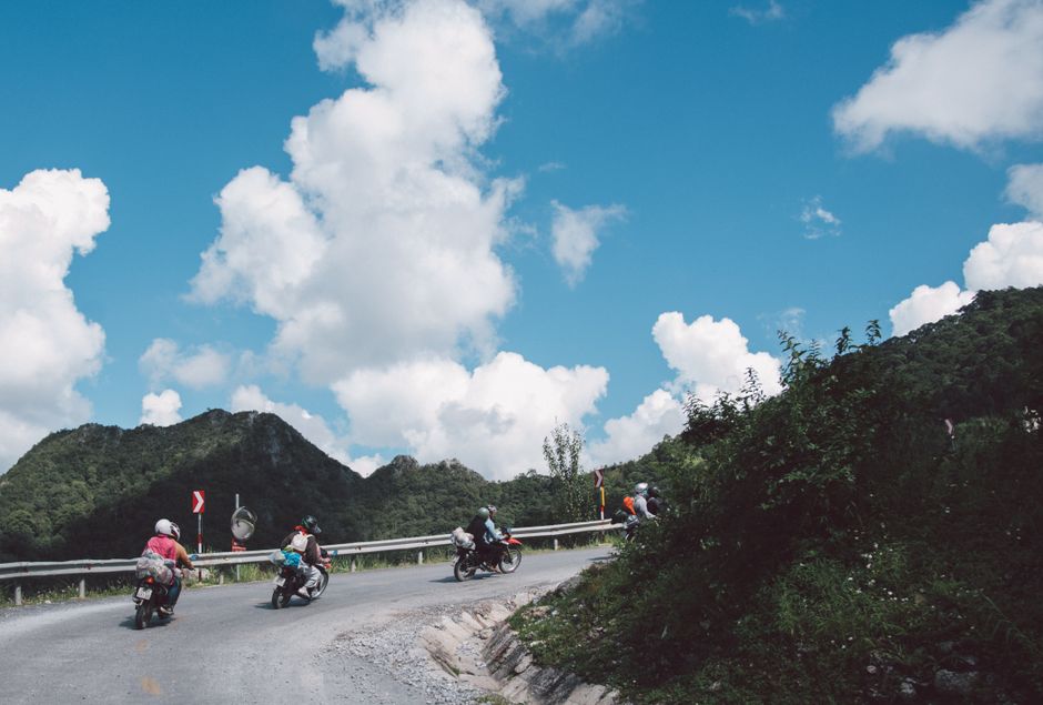 Motorcyclists navigate a winding mountain road under a bright blue sky with fluffy clouds.