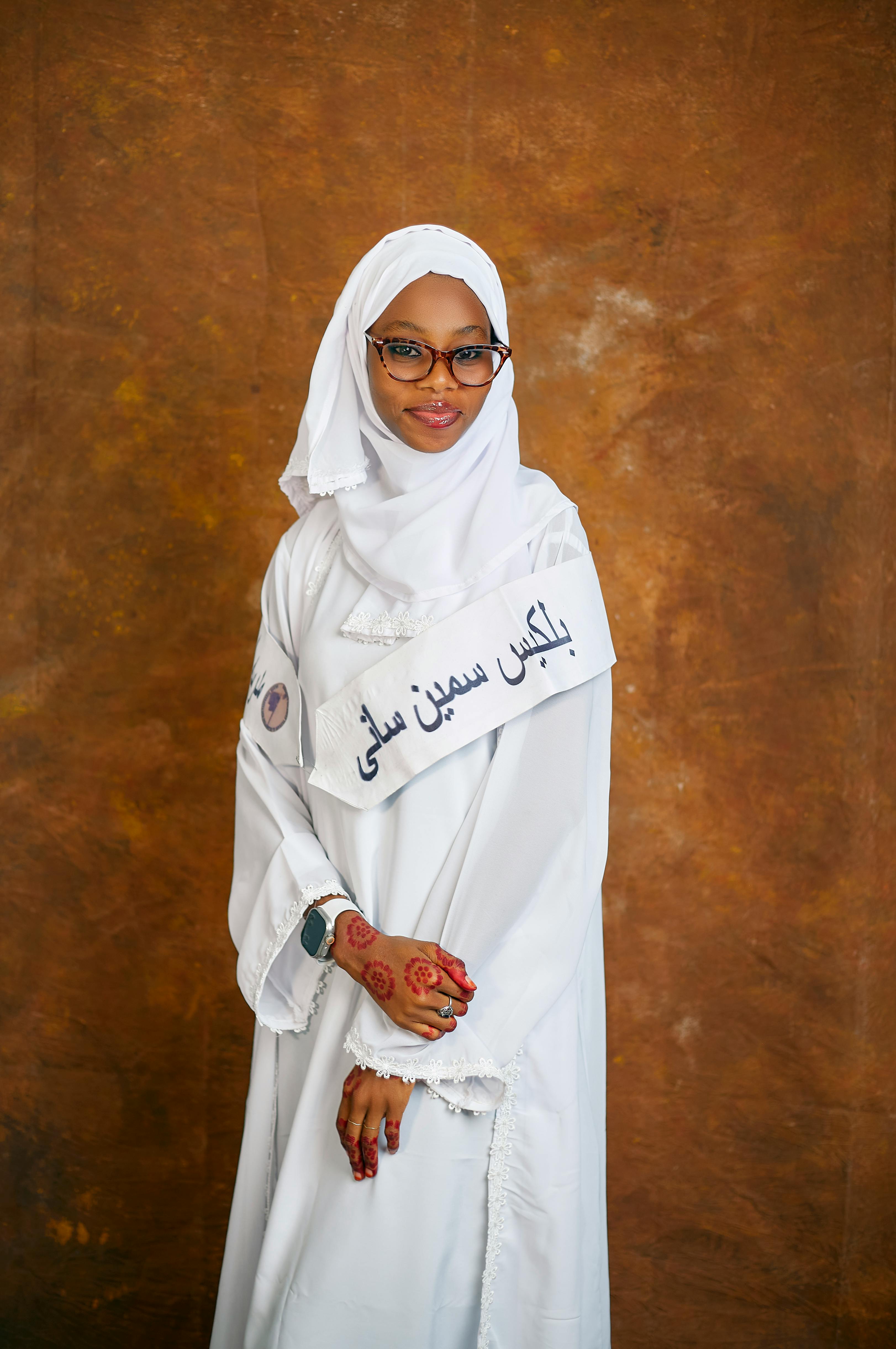 A woman in traditional white attire with henna designs poses against a brown background.