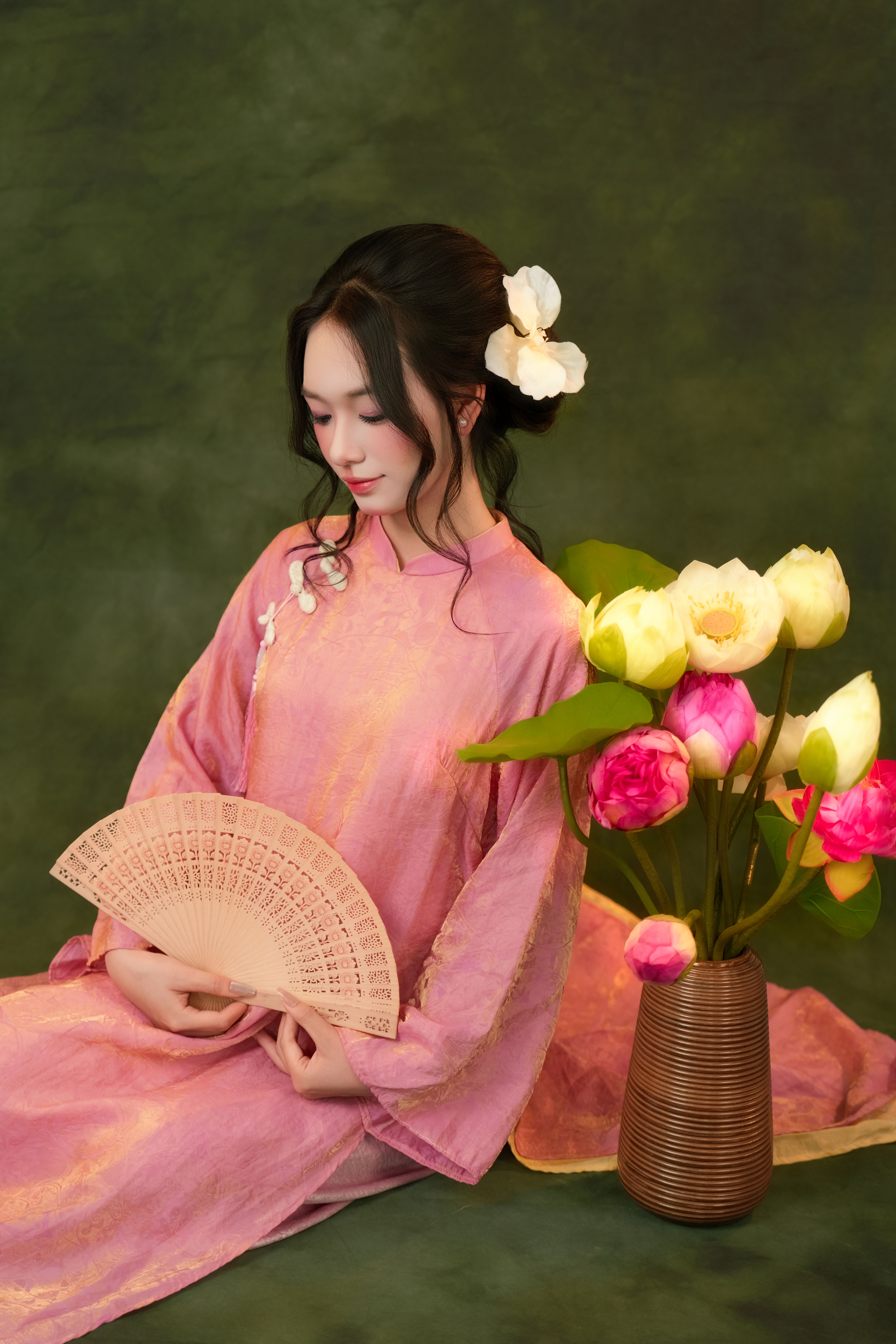 Serene portrait of a woman in a pink dress holding a fan beside a flower vase.