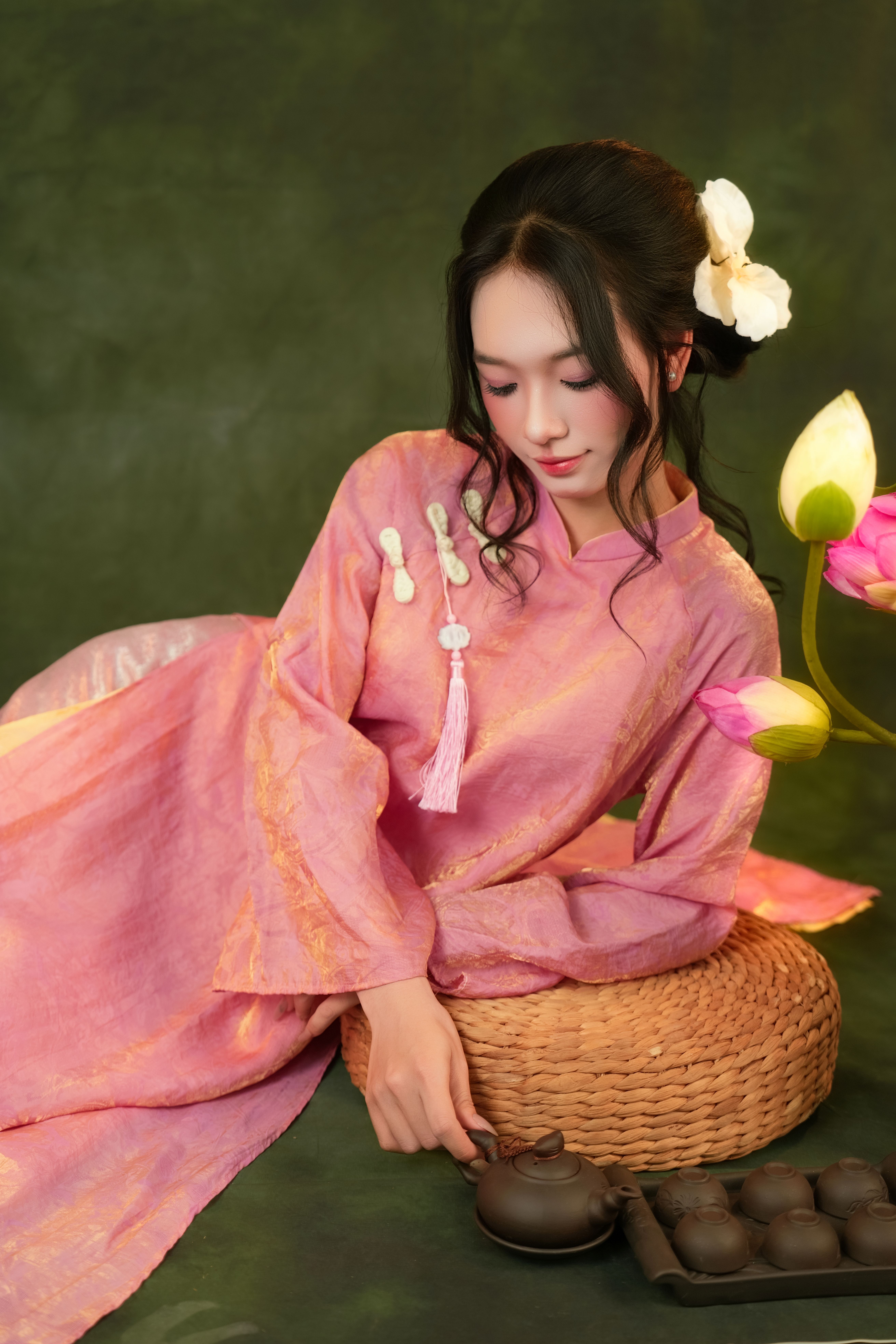 Asian woman in pink traditional dress posing with tea set and lotus flowers.