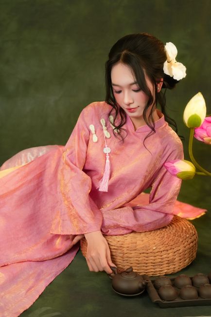 Asian woman in pink traditional dress posing with tea set and lotus flowers.
