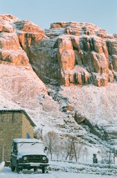 A serene winter scene of snow-covered mountains and a vintage vehicle parked near a rustic building.