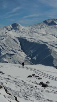 A solitary figure hiking across snow-covered mountainous terrain under a bright sky.