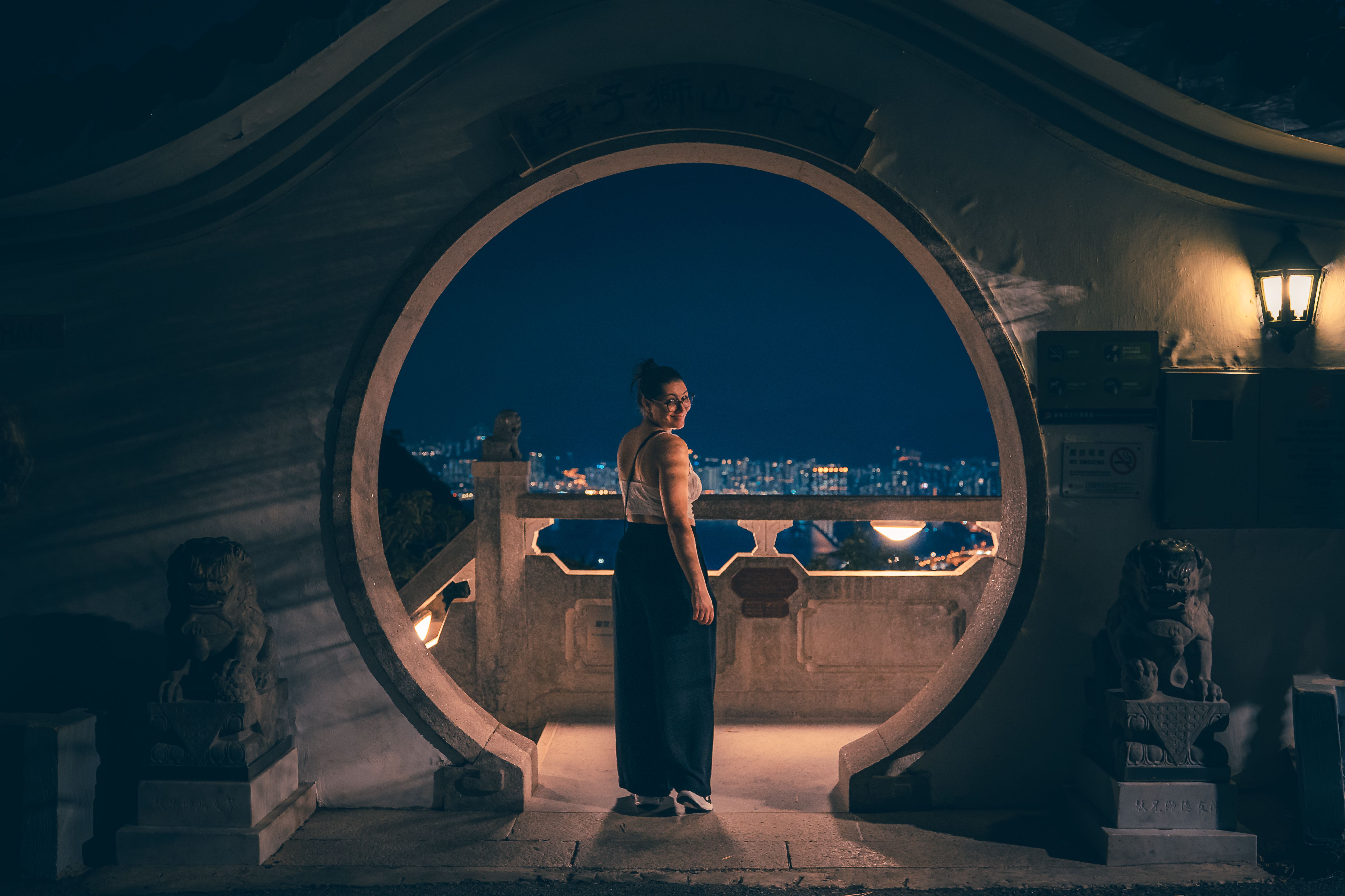 A woman stands by a moon gate, overlooking the city skyline at night on Hong Kong Island.