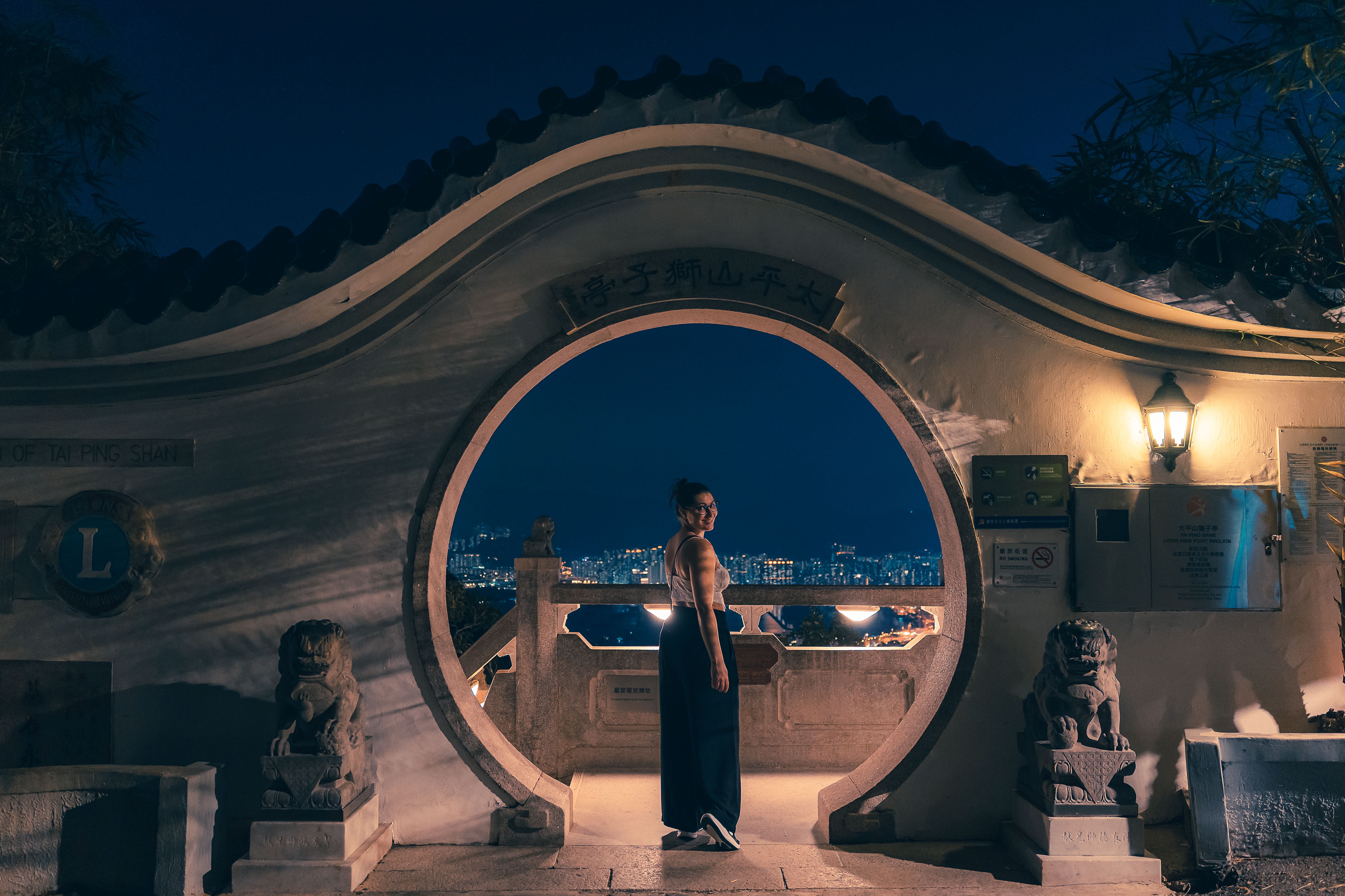 Woman stands at Hong Kong's Tai Ping Shan viewpoint at night, overlooking city lights.