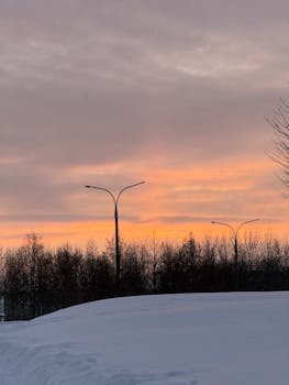 A peaceful winter scene showcasing a vivid sunset over a snowy landscape with trees and streetlights.
