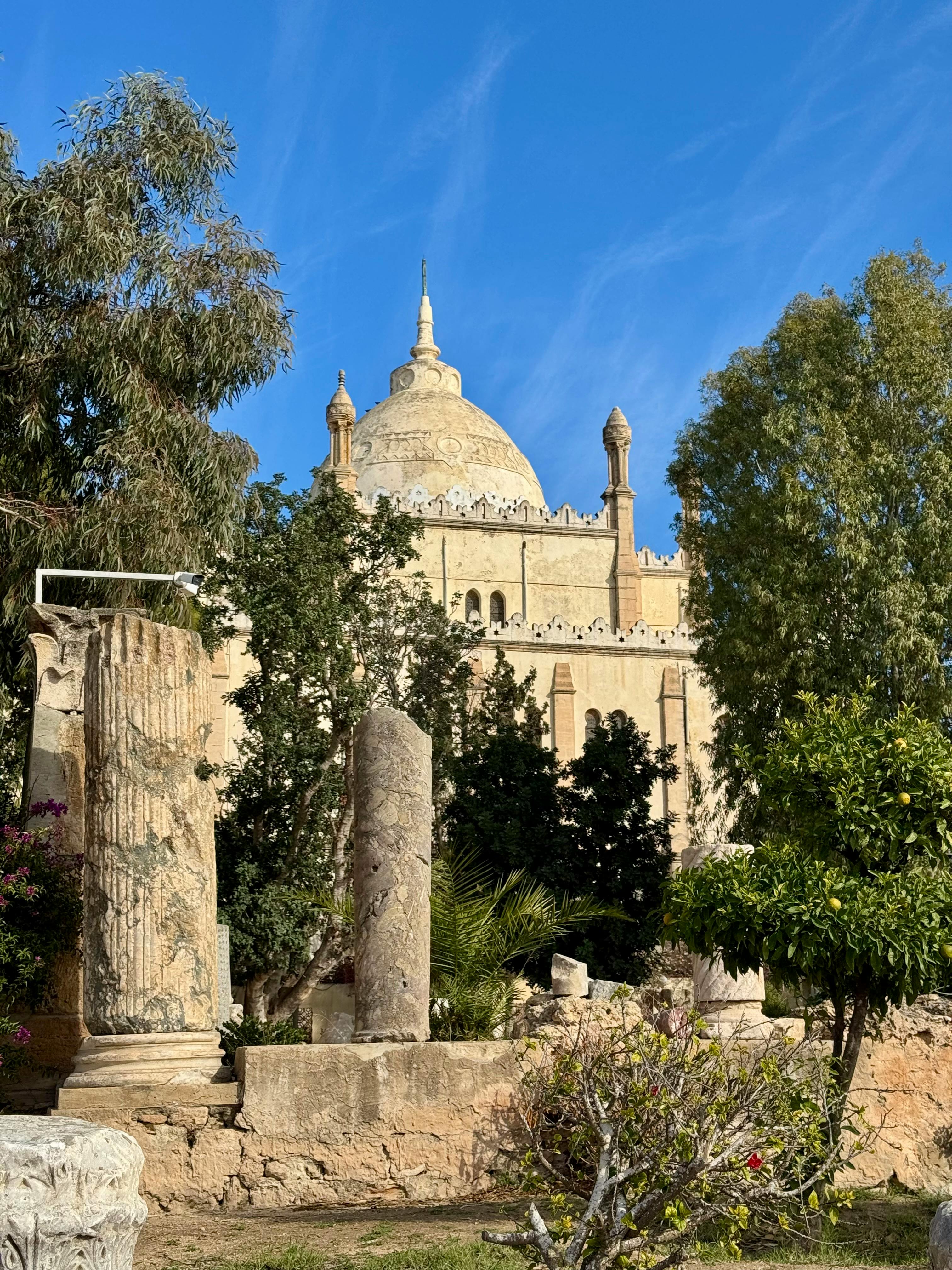 Scenic view of a historic mosque surrounded by ancient ruins and lush greenery under a clear blue sky.