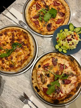 Three delicious pizzas with assorted toppings and a fresh salad on a marble table.