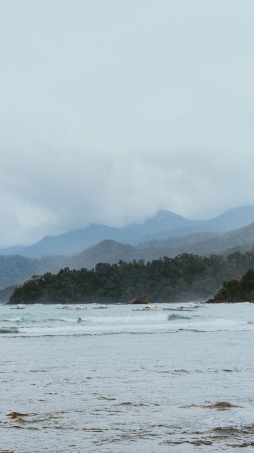Free Serene coastal scene featuring misty mountains and gentle waves under a cloudy sky. Stock Photo