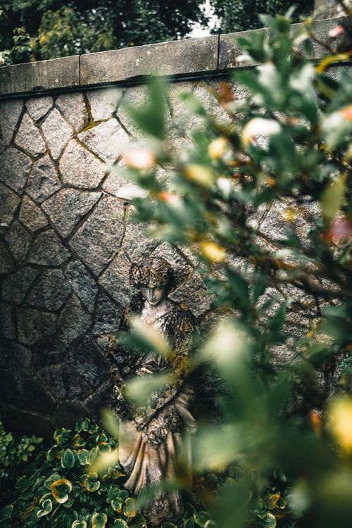 Free A serene garden scene showcasing a weathered stone sculpture amidst lush greenery and a rustic wall. Stock Photo