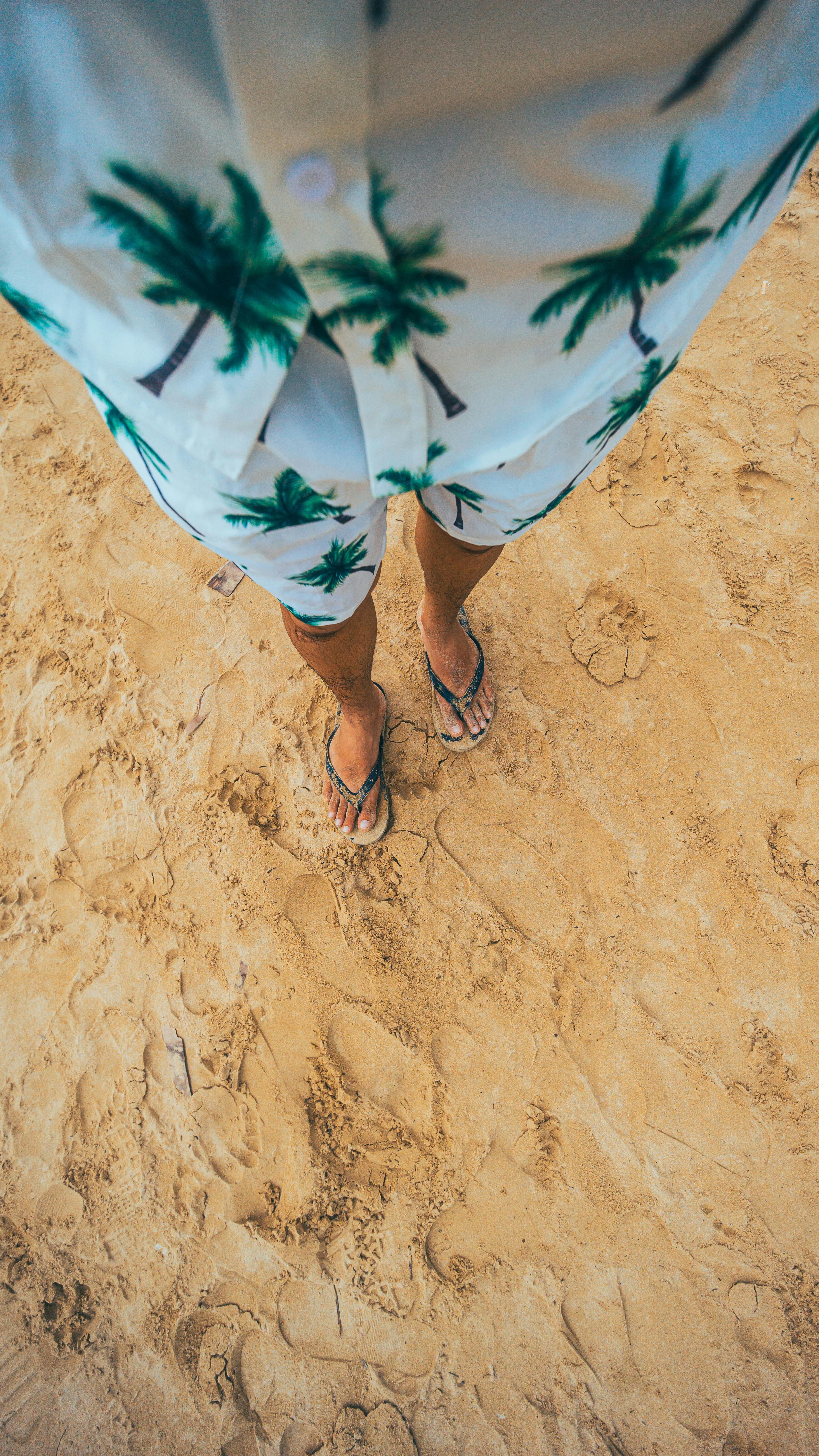 Bezpłatne Osoba ubrana w strój plażowy z odciskiem palm stoi na piaszczystej plaży, przywołując atmosferę letnich wakacji. Zdjęcie z galerii