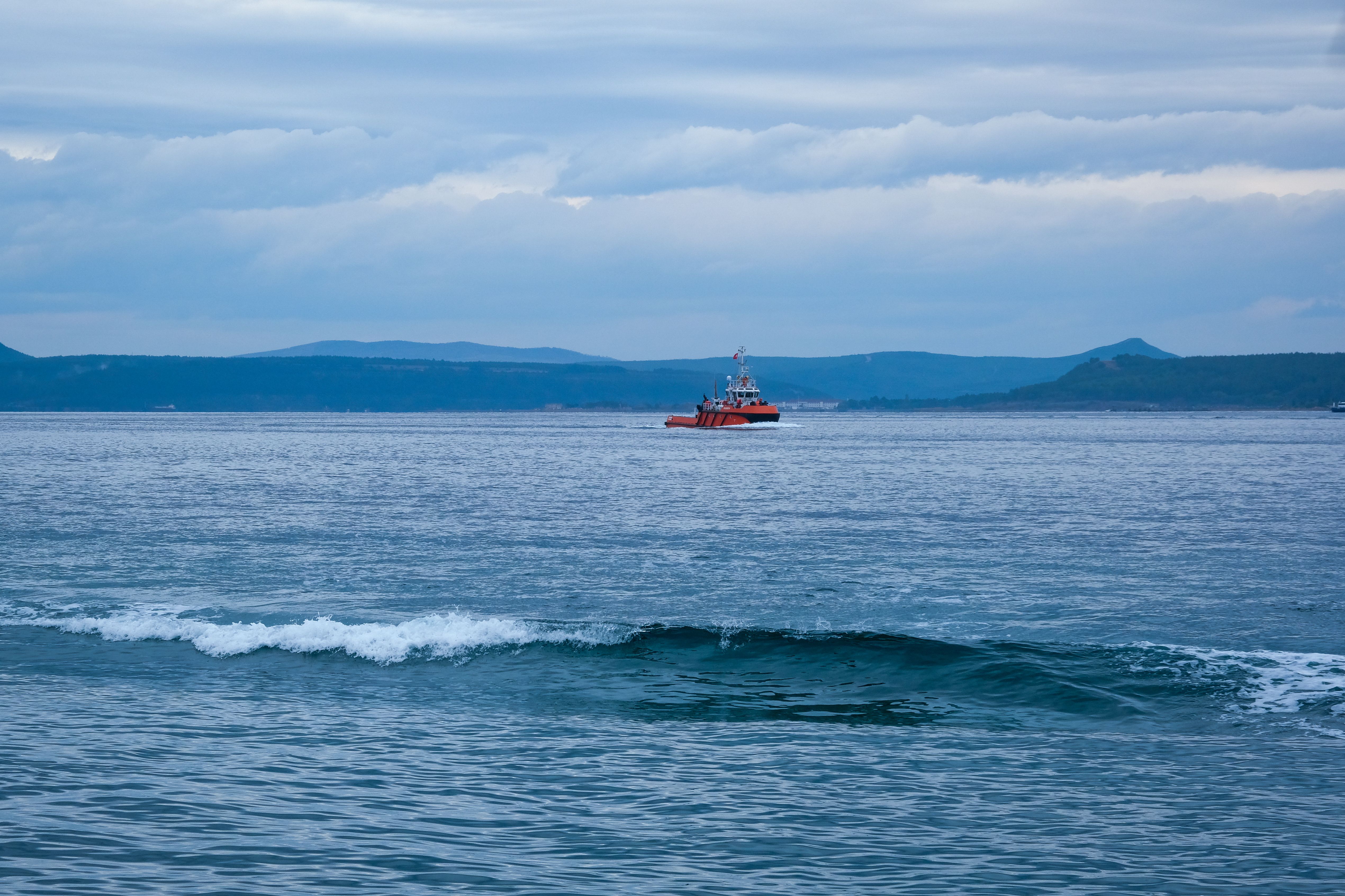 Red tugboat sailing on calm ocean waters with mountains in the background and a cloudy sky.