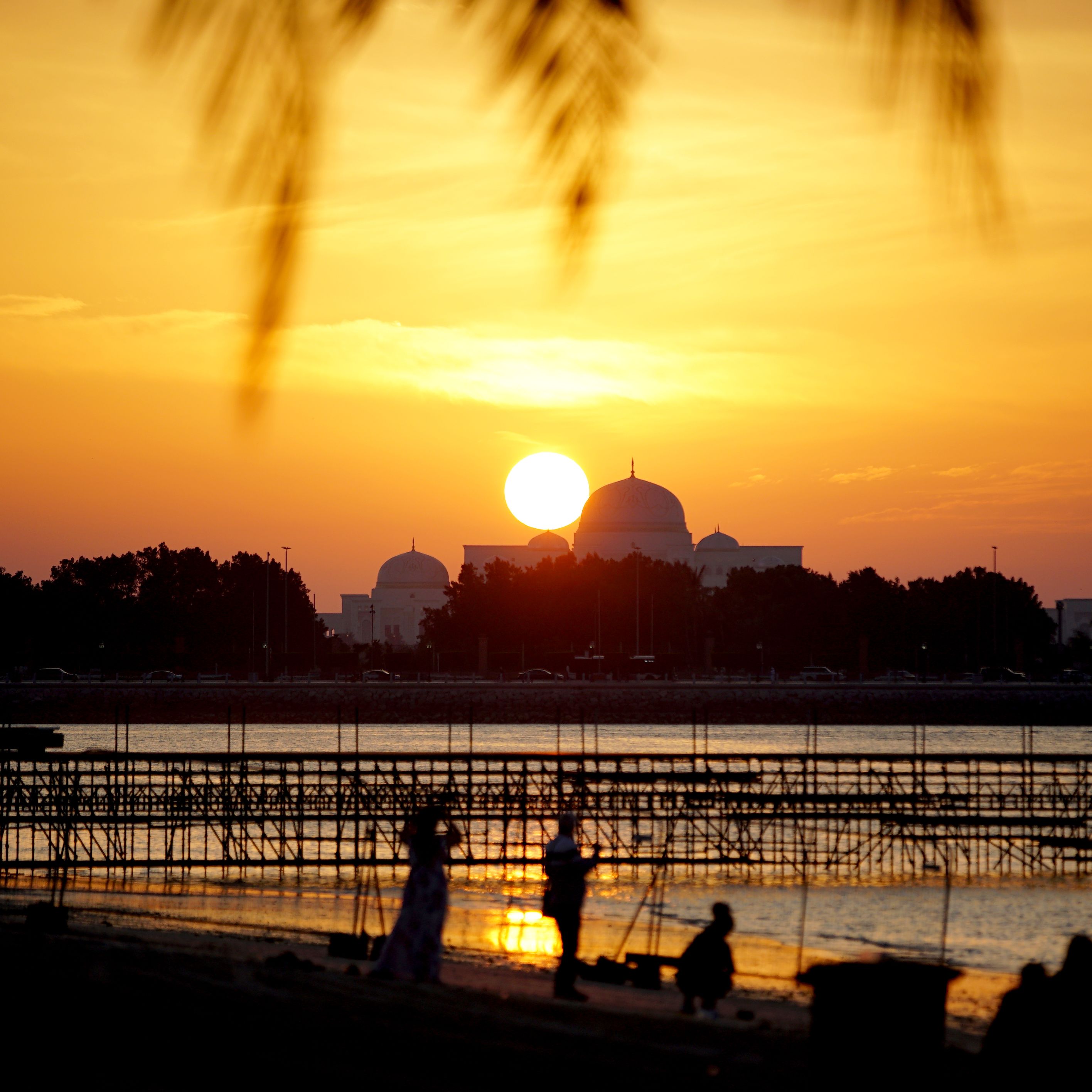 Silhouette of people at sunset by a mosque, casting golden reflections on the water.