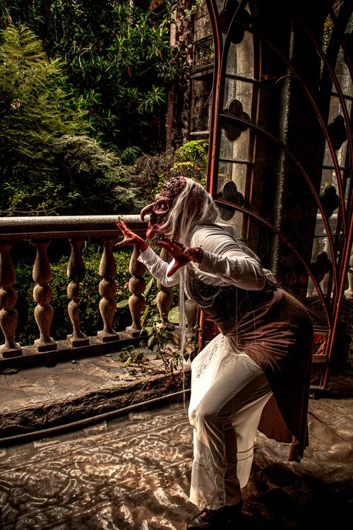 Free A figure dressed as a mystical creature or forest spirit peers off an old balcony enveloped by lush vegetation. Stock Photo